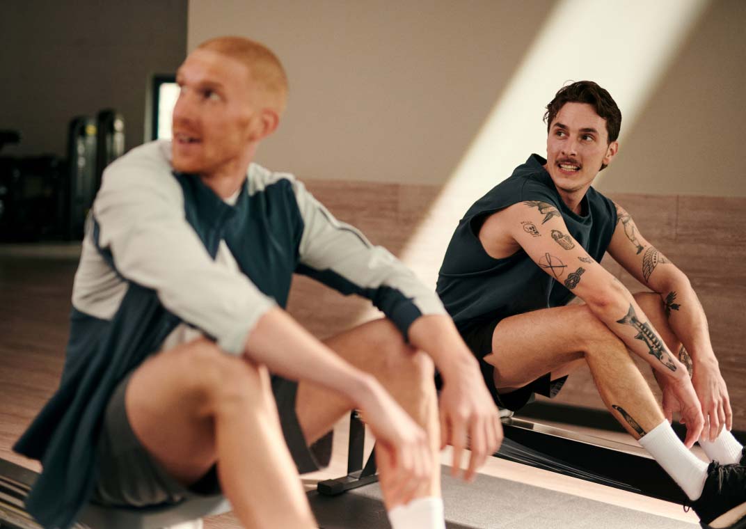 Bild: Zwei Männer sitzen ausgepowert auf Rudergeräten im Cardiobereich. Beide schauen lachend zu einer Person außerhalb des Bildausschnitts.