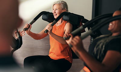Bild: Eine ältere Frau trainiert im Zirkelbereich an einem Gerät mit gepolsterten Bügeln. Sie trägt ein orangenes Shirt und lächelt während der Übungsausführung.