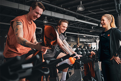 Bild: Zwei Männer sitzen lachend auf Indoor-Cycling-Rädern im Cardiobereich. Neben ihnen steht eine FitX-Trainerin im schwarzen Outfit und unterhält sich mit ihnen.