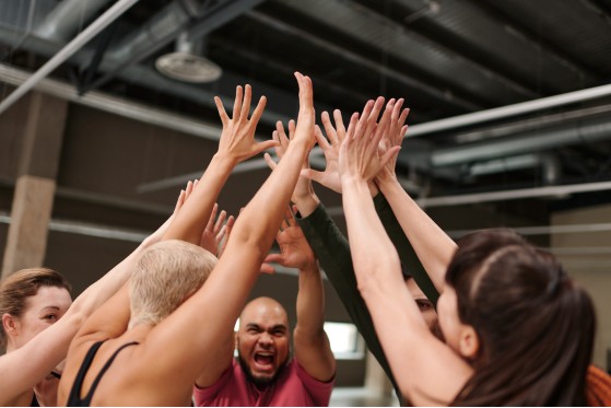 Bild: Eine Gruppe Menschen steht im Kreis. Sie strecken die Arme nach oben für ein gemeinsames High Five. Alle sind motiviert und voller Energie.