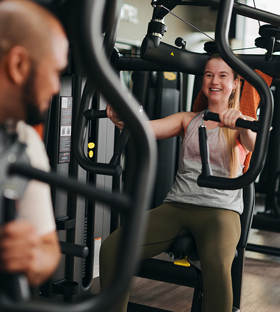 Bild: Eine junge Frau trainiert im Gerätebereich an einer Brustpresse. Sie lächelt einen Mann an, der sich ihr zuwendet.