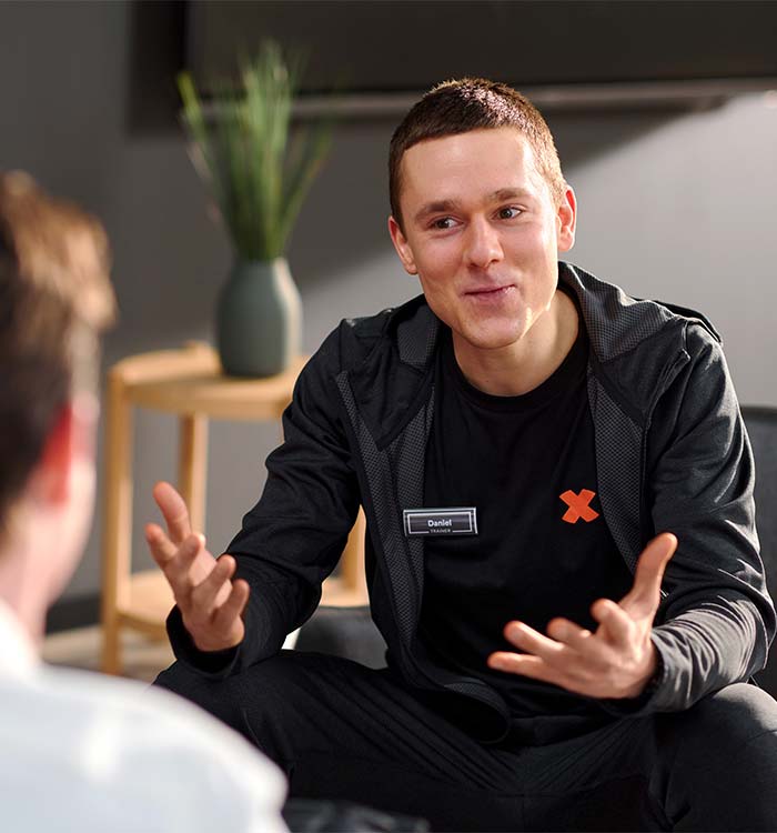 Bild: Ein FitX-Mitarbeiter spricht in einem Beratungsgespräch mit einer weiteren Person. Er lächelt und gestikuliert freundlich mit den Händen.